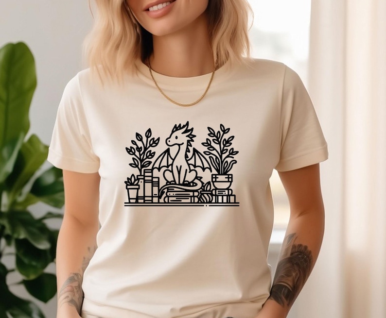 May include: A white t-shirt with a black line drawing of a dragon sitting on a stack of books with plants around it.