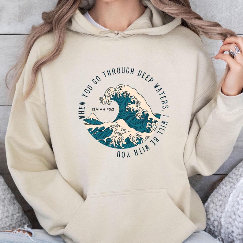 K&ouml;nnte beinhalten: Beigefarbener Hoodie mit einem blauen Wellendesign und dem Text "When you go through deep waters I will be with you" und "Isaiah 43:2".