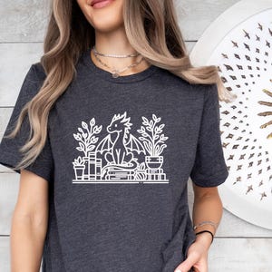 May include: A dark gray t-shirt with a white graphic of a dragon sitting on a stack of books and surrounded by plants. The dragon has wings and a long tail.
