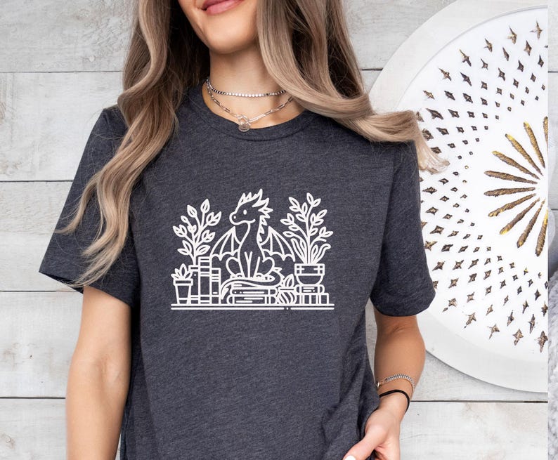 May include: A dark gray t-shirt with a white graphic of a dragon sitting on a stack of books and surrounded by plants. The dragon has wings and a long tail.