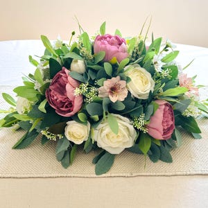May include: A decorative floral arrangement with pink peonies, white roses, and small white flowers, complemented by green foliage. The display is set on a beige fabric, offering a natural and elegant aesthetic.