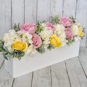 May include: A white rectangular planter overflowing with a variety of artificial flowers, including pink roses, yellow roses, white hydrangeas, and pink peonies.