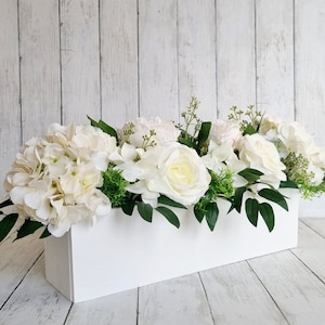 May include: A white rectangular planter filled with a floral arrangement of white hydrangeas, roses, and peonies. The planter is sitting on a white wood background.