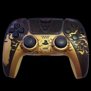 May include: Custom-designed video game controller with a gold and black color scheme. The controller features a silhouette of a bat and bat symbols, with a Joker face on the top. The buttons and joysticks are black.