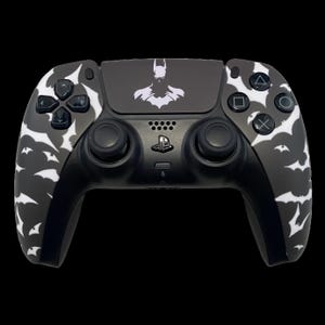 Puede incluir: Un mando de PlayStation 5 en blanco y negro con temática de Batman. El mando presenta un patrón de murciélagos blancos en los laterales y un logotipo de Batman en el panel táctil. Los botones y joysticks son negros.