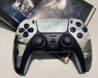 May include: A silver and black wireless game controller featuring a silhouette of a character on the left grip and the signature Lara Croft printed in black on the central touchpad. The controller includes standard black analogue sticks and buttons.