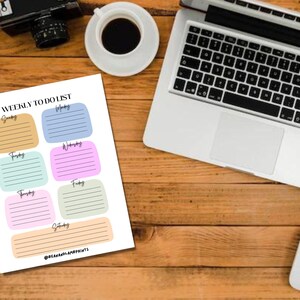 May include: A weekly to-do list on a white paper with colorful boxes for each day of the week. The list is on a wooden desk with a laptop, coffee cup, camera, pen, and a mouse.