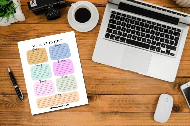 May include: A weekly to-do list on a white paper with colorful boxes for each day of the week. The list is on a wooden desk with a laptop, coffee cup, camera, pen, and a mouse.