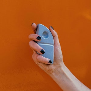 May include: A hand holding a light grey ceramic pipe with a round hole in the top. The pipe is shaped like a fish.