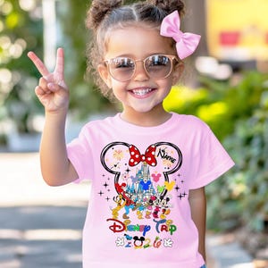K&ouml;nnte beinhalten: Ein rosa T-Shirt mit einer bunten Disney Trip 2023 Grafik mit Mickey Mouse, Minnie Mouse und anderen Charakteren. Das Shirt hat ein Minnie Mouse-Schleifendesign mit dem Wort "Name" darauf.
