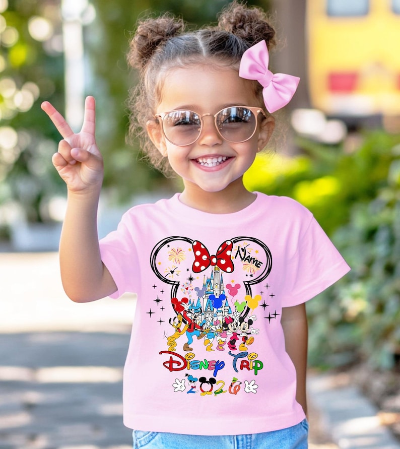 K&ouml;nnte beinhalten: Ein rosa T-Shirt mit einer bunten Disney Trip 2023 Grafik mit Mickey Mouse, Minnie Mouse und anderen Charakteren. Das Shirt hat ein Minnie Mouse-Schleifendesign mit dem Wort "Name" darauf.