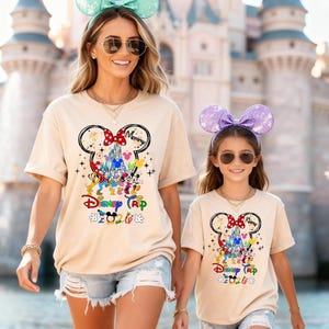 K&ouml;nnte beinhalten: Zwei beige T-Shirts mit einem Mickey Mouse-Design, das ein Schloss, Charaktere und den Text "Disney Trip 2020" zeigt. Die Shirts werden von zwei Personen getragen, die jeweils M&auml;useohren-Stirnb&auml;nder und Sonnenbrillen tragen.