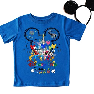 K&ouml;nnte beinhalten: Blaues T-Shirt mit einem farbenfrohen Disney Camp-Motiv, das Micky Maus und Freunde vor einem Schloss zeigt. Das Shirt enth&auml;lt den Text "Disney Trip 2020" und einen Platz f&uuml;r einen Namen. Ein Paar schwarze Micky Maus-Ohren ist im Hintergrund.