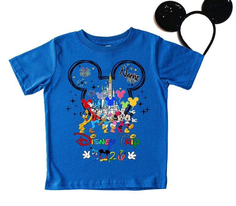 K&ouml;nnte beinhalten: Blaues T-Shirt mit einem farbenfrohen Disney Camp-Motiv, das Micky Maus und Freunde vor einem Schloss zeigt. Das Shirt enth&auml;lt den Text "Disney Trip 2020" und einen Platz f&uuml;r einen Namen. Ein Paar schwarze Micky Maus-Ohren ist im Hintergrund.