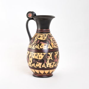 Op de afbeelding: Een zwarte en gele keramische kan met een handvat en een decoratieve bovenkant. De kan is versierd met een zwart en rood ontwerp van leeuwen en andere mythische wezens.