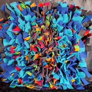 May include: A colorful dog bed made of fleece with blue, turquoise, gray, orange, yellow, and red fabric strips. The bed is designed to be stimulating for dogs and encourages them to play.