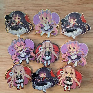 May include: Nine enamel pins featuring anime-style characters. The characters are wearing dresses and have pink and black hair. The pins are gold-colored and have a heart-shaped design.