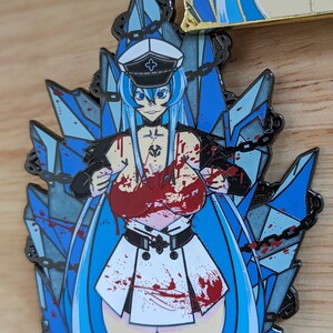 May include: A blue and white enamel pin featuring a female anime character with long blue hair, wearing a white and black military uniform with a red cross on the collar. The character is surrounded by blue ice crystals and chains. The character is covered in red blood splatters.