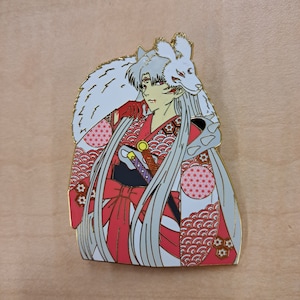 Puede incluir: Un pin de esmalte dorado con un personaje de anime de pelo blanco que lleva un kimono rojo y blanco y una cabeza de lobo blanco. El personaje tiene ojos rojos y sostiene una espada. El pin tiene un diseño detallado con patrones intrincados.