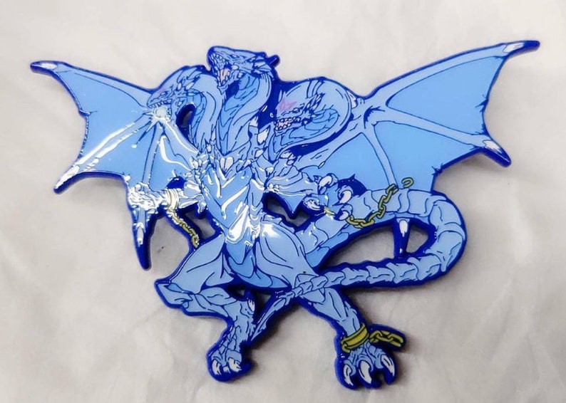 May include: A blue enamel pin depicting a three-headed dragon with chains around its legs and wings. The dragon is standing with its wings outstretched.