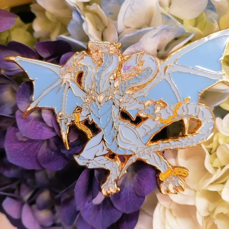 May include: A blue and gold enamel pin featuring a three-headed dragon with wings outstretched. The dragon is depicted in a stylised, fantasy-inspired design.