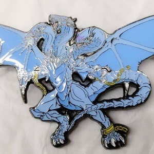 May include: A blue and silver metal pin of a dragon with its wings spread out. The dragon is chained at the wrists and ankles.