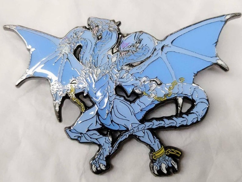 May include: A blue and silver metal pin of a dragon with its wings spread out. The dragon is chained at the wrists and ankles.