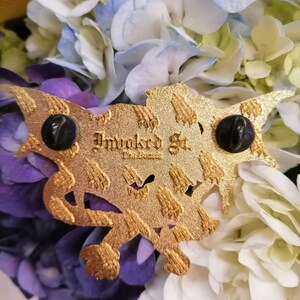 May include: Gold-coloured metal pin featuring a detailed dragon design. The back of the pin has a black rubber clutch and the text "Invoked St.  The Unseen" printed in black.