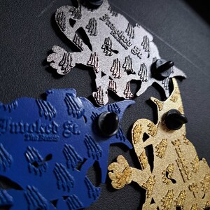 May include: Three enamel pins featuring a stylised lizard design. The pins are in silver, gold, and blue with a textured finish. Each pin has the text "Fraudulent Et. The Brand" printed on it.