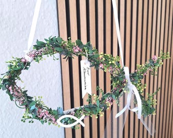 Puede incluir: Una corona con forma de pez hecha de follaje verde y pequeñas flores rosas, suspendida por cintas blancas. Una etiqueta blanca cuelga con el texto "Zur Kommunion" en negro, y un símbolo de pez blanco está sujeto en la parte inferior.