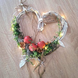 Könnte beinhalten: Ein herzförmiger Kranz aus weißen Zweigen, verziert mit drei roten Äpfeln, grünem Blattwerk, Moos und kleinen weißen Lichterketten. Ein kleines braunes Schild mit der Aufschrift "Hallo Herbst" ist mit Juteschnur und metallenen Blattanhängern befestigt.