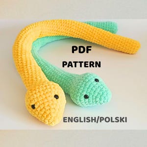 Puede incluir: Dos juguetes de serpiente de ganchillo, uno amarillo y otro verde, con ojos de botón negros. El texto "PDF PATTERN" y "ENGLISH/POLSKI" es visible. Las serpientes están hechas de un hilo suave y texturizado.