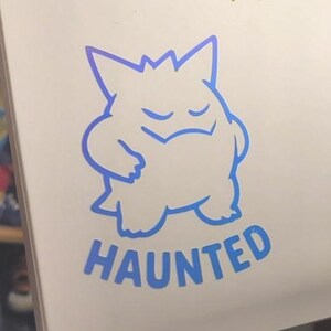 May include: A white rectangular surface featuring three distinct decals. At the top, yellow text reads DOOR TOUCHER above a car door icon. Below, a yellow line-art illustration of a person wearing a headset with the text PIT CREW. At the bottom, a blue line-art illustration of a Gengar character with the text HAUNTED.