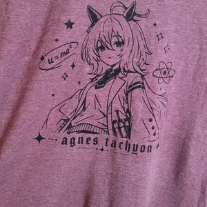 May include: A heathered mauve t-shirt featuring a black line art graphic of an anime character with animal ears, wearing a lab coat. The design includes the text "u=ma&sup2;" and "agnes tachyon," along with small star, sparkle, and atom symbols.
