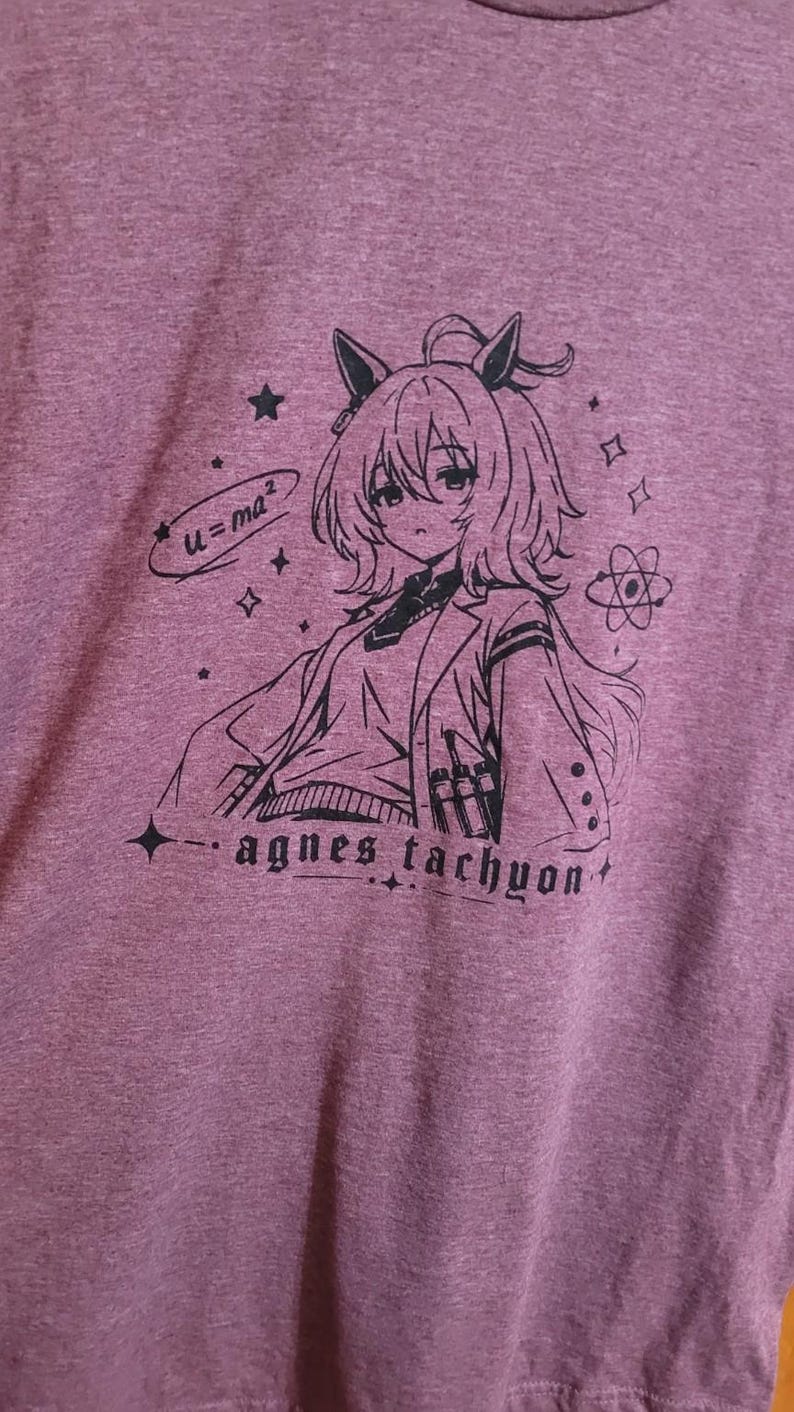May include: A heathered mauve t-shirt featuring a black line art graphic of an anime character with animal ears, wearing a lab coat. The design includes the text "u=ma&sup2;" and "agnes tachyon," along with small star, sparkle, and atom symbols.