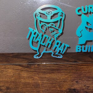 May include: Two teal-colored cutouts on a wooden surface. One cutout features a rat holding a steering wheel with the text "TRACK RAT". The other cutout is a cartoon character with the text "CUR BUILD".