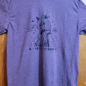 May include: A heather purple short-sleeve t-shirt featuring a black line art graphic of an anime-style character with cat ears. The design includes the text "u=ma&sup2;", an atom symbol, stars, and the name "agnes tachyon" printed in a stylized font.