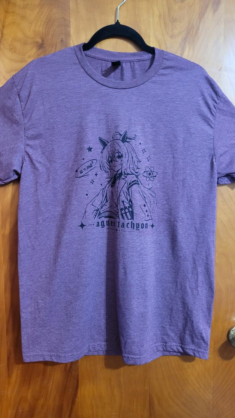 May include: A heather purple short-sleeve t-shirt featuring a black line art graphic of an anime-style character with cat ears. The design includes the text "u=ma&sup2;", an atom symbol, stars, and the name "agnes tachyon" printed in a stylized font.