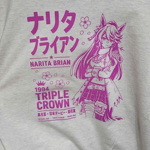 May include: A heathered beige t-shirt featuring a magenta graphic print of an anime-style character with horse ears. The text reads Narita Brian, 1994 Triple Crown, and includes Japanese characters along with Satsuki Sho, Tokyo Yushun, and Kikuka Sho.