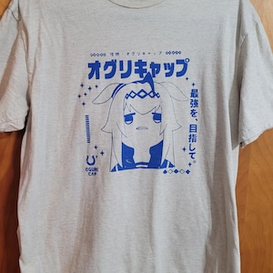 May include: A light grey heathered t-shirt featuring a blue graphic print of an anime-style character with long hair and ears. The design includes Japanese text, the words OGURI CAP, and small star accents centred on the front of the garment.