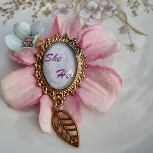 Könnte beinhalten: Eine goldfarbene Brosche mit einem weißen Kamee, auf dem die Worte "She Her" in lila Schrift stehen. Der Kamee ist von einem dekorativen goldenen Rahmen umgeben und hängt an einem goldenen Blattanhänger.