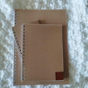 May include: Three spiral-bound notebooks in varying sizes, featuring a textured tan cover. The notebooks are stacked, with the largest at the bottom and the smallest on top. A small, rectangular label is affixed to the bottom notebook.