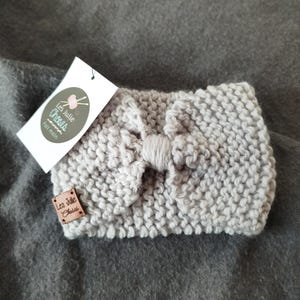 May include: A light gray knitted headband with a large bow in the center. The headband has a brown leather tag with the text "Les Julie Choses" and "Fait main".