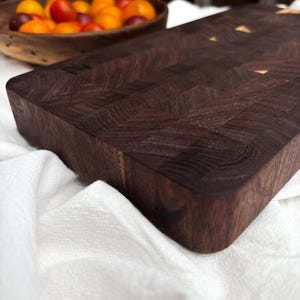 May include: Dark brown end-grain chopping board with a rectangular shape. The board has a mosaic-like pattern and rounded edges. A wooden bowl with orange and red tomatoes is in the background.