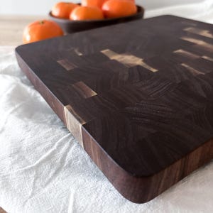 Puede incluir: Una tabla de cortar de madera marrón oscuro con forma rectangular y un patrón similar a un mosaico. La tabla tiene una superficie lisa y pulida con detalles de madera más claros. En el fondo, un cuenco de fruta naranja descansa sobre un paño blanco.