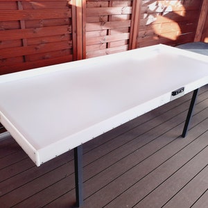 Peut inclure: Table rectangulaire blanche avec des pieds noirs. La table a une surface lisse et est placée devant un fond en bois. Ses dimensions sont d'environ 152 cm de long et 76 cm de large.