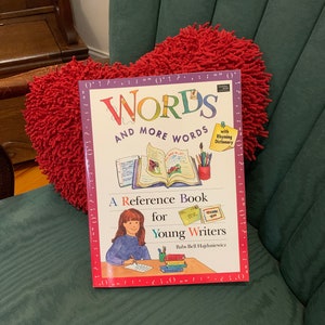 May include: A red book titled "Words and More Words" with a rhyming dictionary. The book is a reference book for young writers. The cover features a girl writing at a desk with pencils and paper.