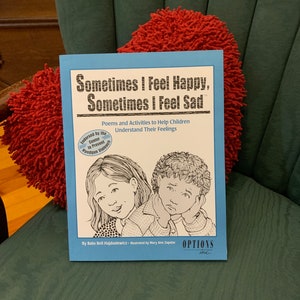May include: A blue book titled "Sometimes I Feel Happy, Sometimes I Feel Sad" with a red heart-shaped pillow in the background. The book is about poems and activities to help children understand their feelings. It is endorsed by the Center to Prevent Handgun Violence.