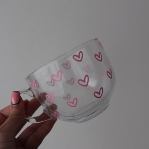 K&ouml;nnte beinhalten: Eine klare Glas-Tasse mit einem Muster aus rosa Herzen. Die Tasse hat einen Henkel und wird in einer Hand gehalten.