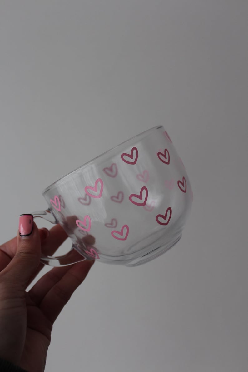 K&ouml;nnte beinhalten: Eine klare Glas-Tasse mit einem Muster aus rosa Herzen. Die Tasse hat einen Henkel und wird in einer Hand gehalten.
